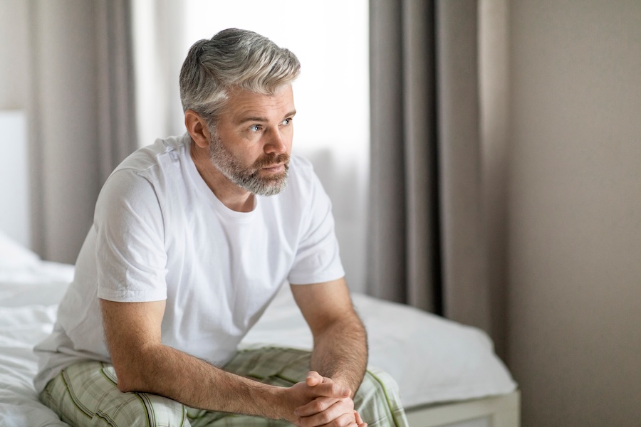 Pensive thoughtful unhappy handsome grey-haired bearded mature caucasian man wearing pajamas sitting on bed at home, looking at copy space, feeling lonely, going through divorce