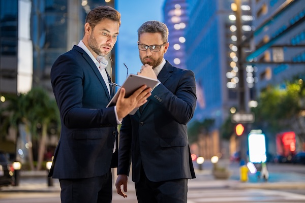 Business men in night Ney York. Business people discuss a project. The two business teams men team using tablet laptop outdoor. Businessmen looking tablet with business success in night city.
